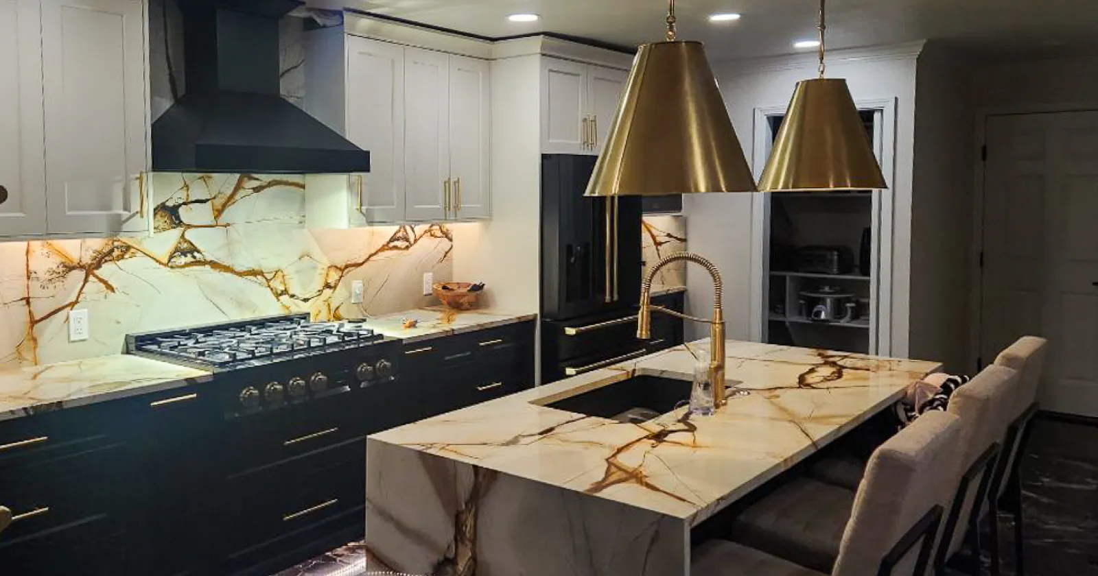 Luxurious modern kitchen with bold black and white cabinetry, striking marble waterfall island and backsplash with dramatic veining. Features include brass pendant lights, black tile flooring, double wall ovens, and a matte black refrigerator and range for a sleek, high-end look.