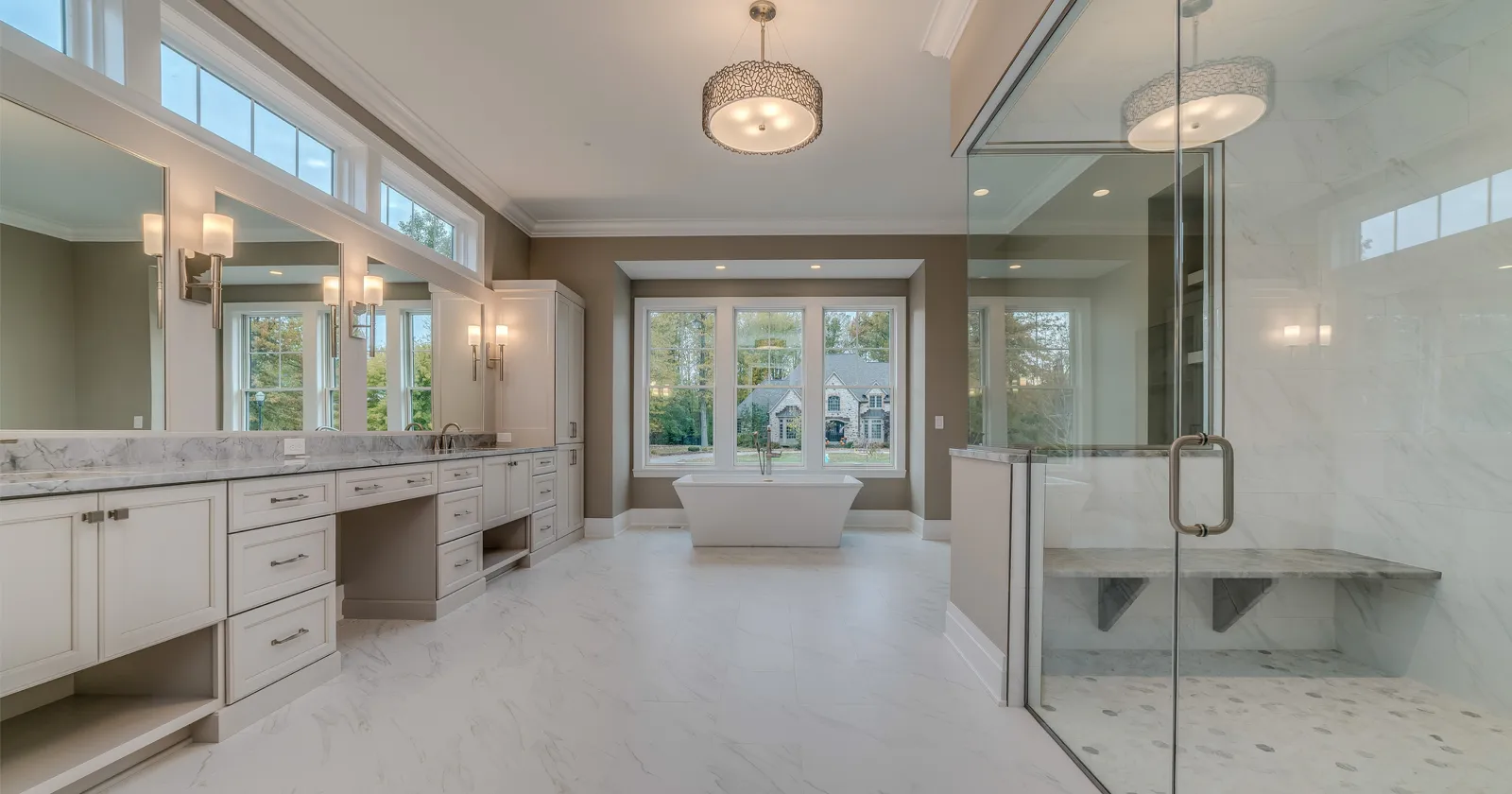 Spacious luxury bathroom featuring a freestanding soaking tub, large glass-enclosed shower with a built-in bench, dual vanities with marble countertops, and expansive windows providing natural light and views of a landscaped yard.