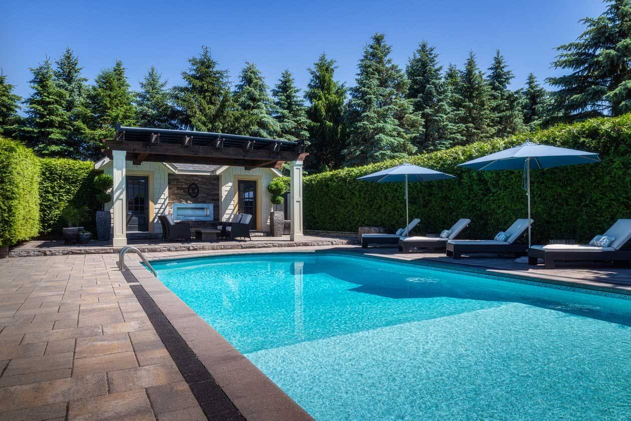 A tranquil outdoor pool area featuring clear blue water, comfortable lounge chairs with umbrellas, and a cozy pergola seating area, all surrounded by lush greenery.