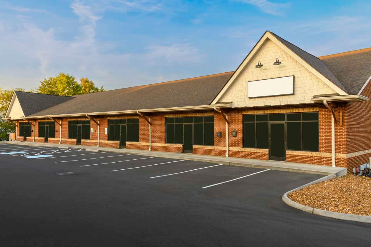A office complex with a brick exterior and large windows covered with black shutters. The parking lot in front is empty with lined parking spaces, and the sky is clear and blue.