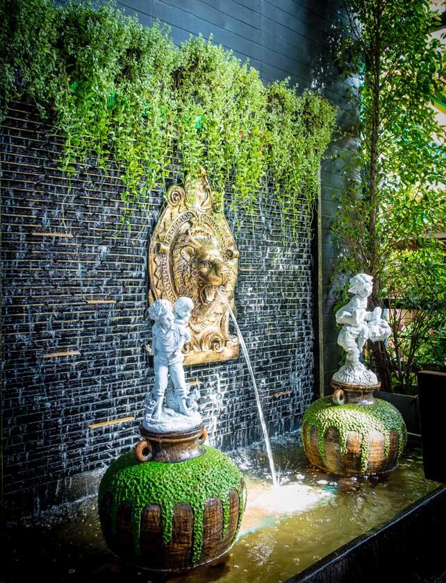 A decorative garden water fountain featuring a lion sculpture with water flowing from its mouth, flanked by two statues of children. The fountain is surrounded by greenery and plants.